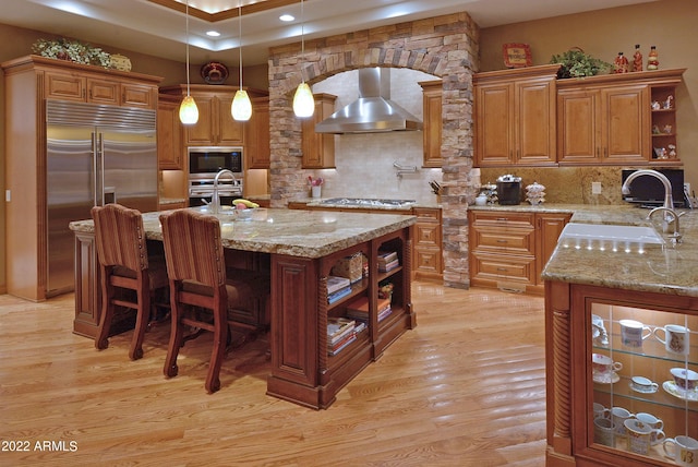 kitchen featuring built in appliances, wall chimney exhaust hood, a center island with sink, and light stone countertops