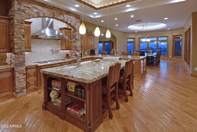 kitchen with backsplash, wall chimney exhaust hood, a large island with sink, pendant lighting, and stainless steel gas stovetop