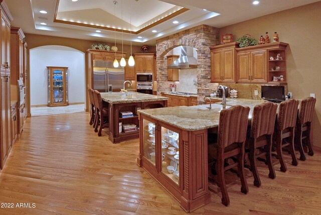 kitchen with light stone counters, wall chimney exhaust hood, built in appliances, decorative light fixtures, and a center island with sink