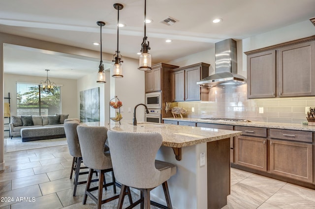 kitchen with backsplash, appliances with stainless steel finishes, a chandelier, wall chimney range hood, and a breakfast bar area