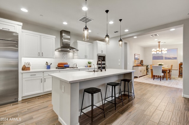 kitchen featuring wall chimney exhaust hood, pendant lighting, stainless steel appliances, an island with sink, and white cabinets