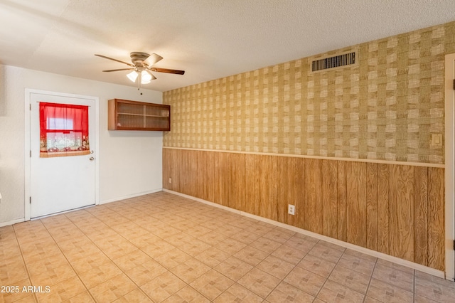 empty room featuring a textured ceiling, ceiling fan, wood walls, visible vents, and wainscoting