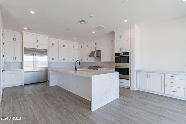 kitchen featuring light stone countertops, appliances with stainless steel finishes, backsplash, a center island with sink, and white cabinetry