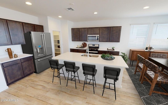 kitchen with a breakfast bar area, an island with sink, dark brown cabinets, light hardwood / wood-style floors, and stainless steel appliances