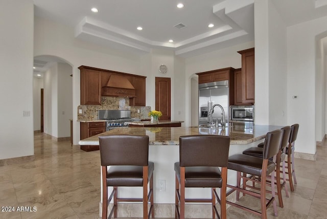 kitchen with backsplash, a tray ceiling, premium range hood, an island with sink, and built in appliances