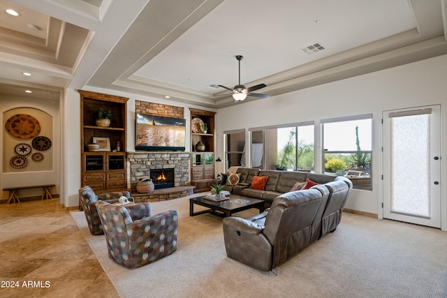 living room featuring a stone fireplace, built in shelves, a tray ceiling, and ceiling fan