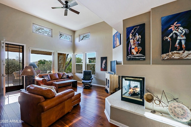 living area with ceiling fan, baseboards, a multi sided fireplace, a high ceiling, and dark wood-style floors