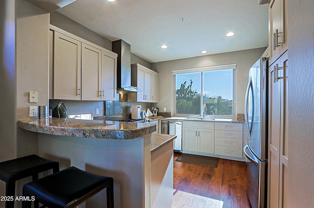 kitchen featuring dark wood finished floors, a peninsula, a sink, appliances with stainless steel finishes, and wall chimney exhaust hood