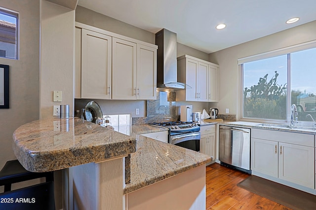 kitchen with a peninsula, a sink, stainless steel appliances, wall chimney range hood, and light wood-type flooring