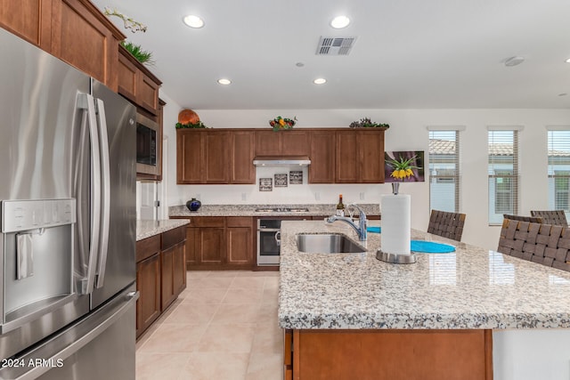 kitchen with an island with sink, sink, light stone countertops, light tile patterned floors, and appliances with stainless steel finishes