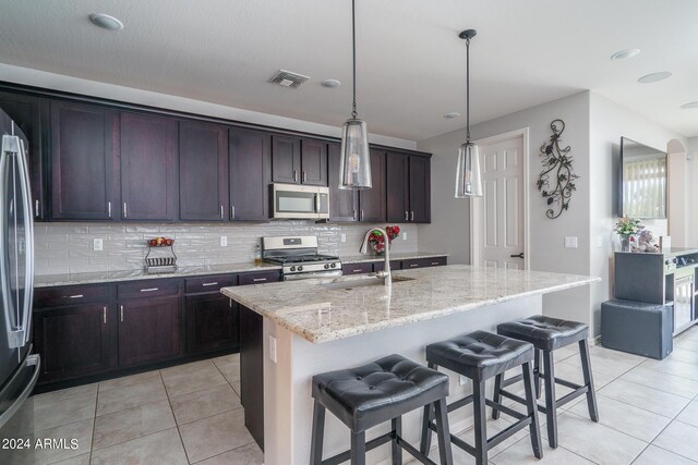 kitchen with an island with sink, sink, stainless steel appliances, light stone countertops, and a kitchen bar