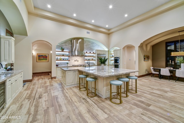 kitchen featuring a center island with sink, light stone counters, wall chimney range hood, and light hardwood / wood-style floors