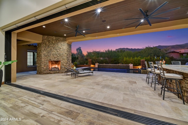 patio terrace at dusk with an outdoor stone fireplace, an outdoor bar, and ceiling fan