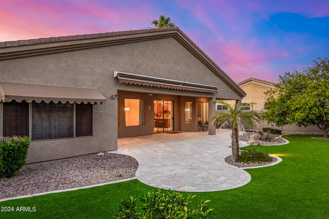 back house at dusk featuring a patio and a lawn