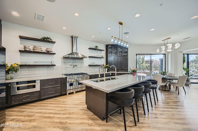 kitchen with open shelves, a sink, light wood-style floors, wall chimney exhaust hood, and appliances with stainless steel finishes