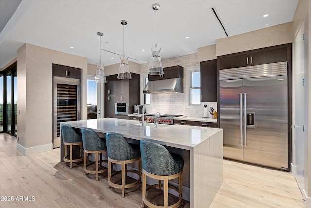 kitchen with light wood finished floors, light stone counters, a kitchen island with sink, stainless steel appliances, and wall chimney range hood