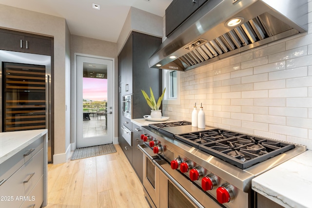 kitchen with tasteful backsplash, wall chimney range hood, light stone countertops, double oven range, and light wood-style floors