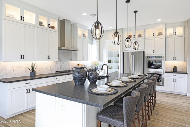 kitchen with stainless steel appliances, white cabinetry, a center island with sink, and wall chimney range hood