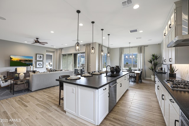 kitchen featuring wall chimney range hood, sink, white cabinetry, a center island with sink, and decorative light fixtures