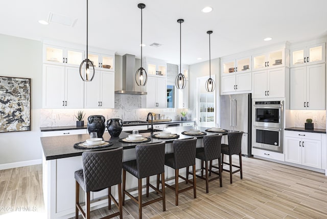 kitchen featuring white cabinetry, hanging light fixtures, stainless steel appliances, a kitchen island with sink, and wall chimney range hood