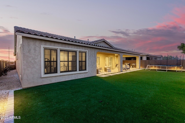 back of house at dusk featuring a lawn, a patio, a tile roof, a trampoline, and stucco siding