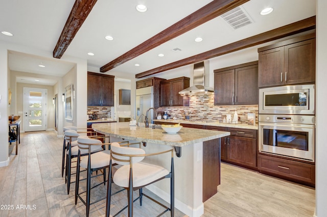 kitchen with a kitchen island with sink, wall chimney range hood, light stone counters, a breakfast bar, and beamed ceiling