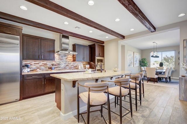 kitchen with tasteful backsplash, wall chimney range hood, built in appliances, a kitchen island with sink, and light stone countertops