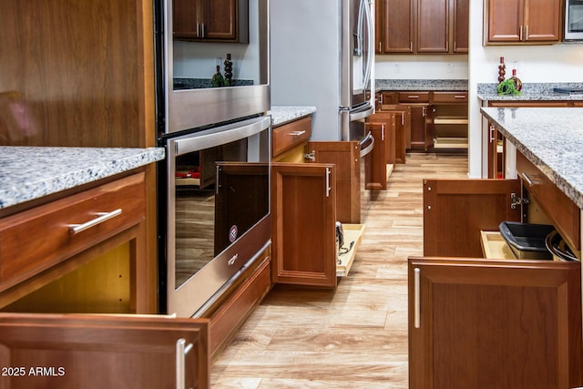 kitchen with stainless steel appliances, light wood-type flooring, brown cabinets, and light stone countertops