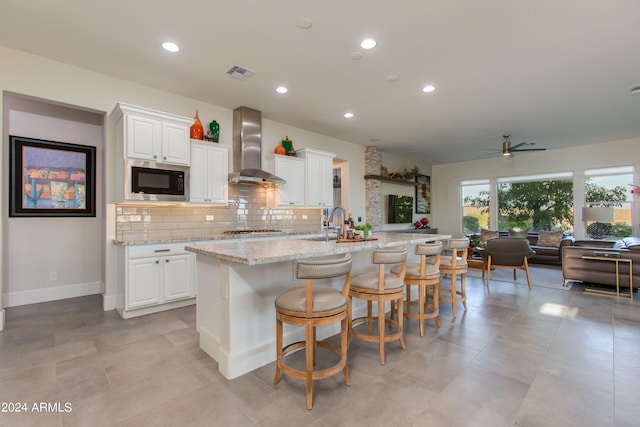 kitchen with wall chimney exhaust hood, white cabinets, an island with sink, and ceiling fan