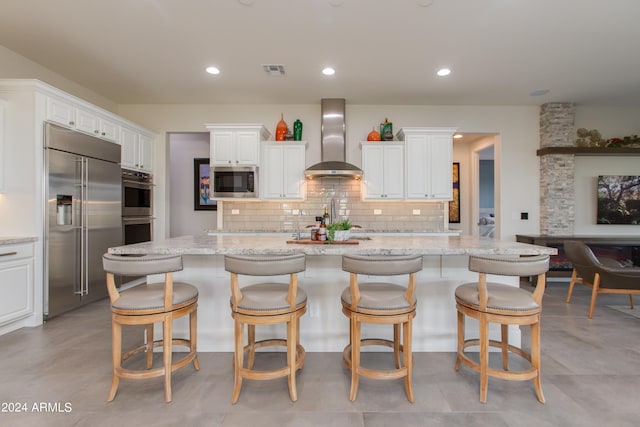 kitchen with an island with sink, white cabinetry, wall chimney range hood, and built in appliances