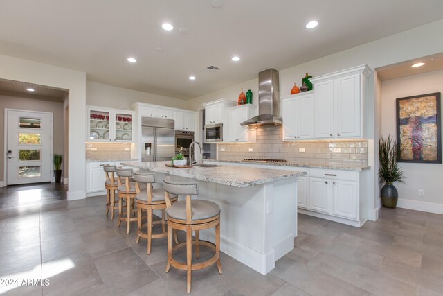 kitchen with decorative backsplash, white cabinetry, wall chimney exhaust hood, stainless steel appliances, and a kitchen island with sink