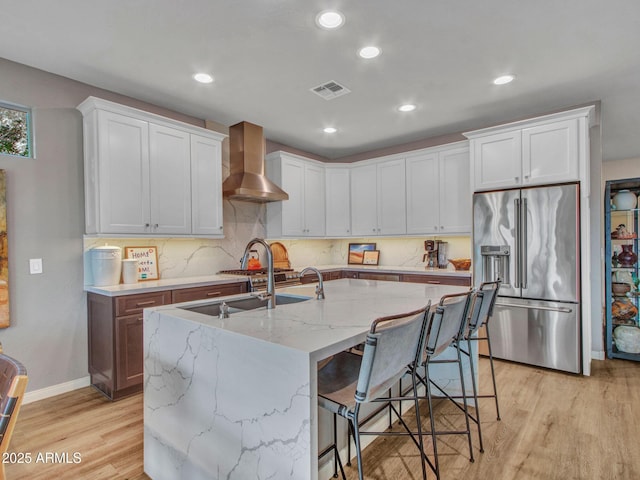 kitchen featuring high quality fridge, white cabinetry, an island with sink, sink, and wall chimney exhaust hood