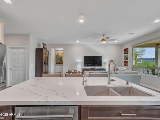 kitchen with sink, light stone counters, a center island with sink, stainless steel fridge, and ceiling fan