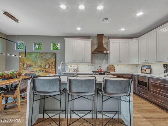 kitchen with white cabinetry, wall chimney exhaust hood, a kitchen breakfast bar, and light wood-type flooring