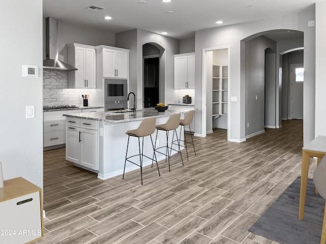 kitchen with wall chimney exhaust hood, white cabinets, light hardwood / wood-style flooring, and a kitchen island with sink