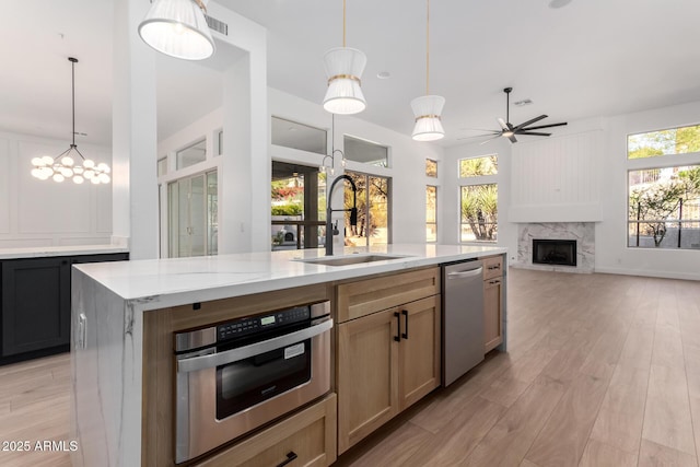 kitchen with appliances with stainless steel finishes, a sink, ceiling fan with notable chandelier, a high end fireplace, and light wood-type flooring