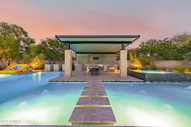 pool at dusk featuring an outdoor kitchen, an in ground hot tub, fence, a fenced in pool, and a patio area