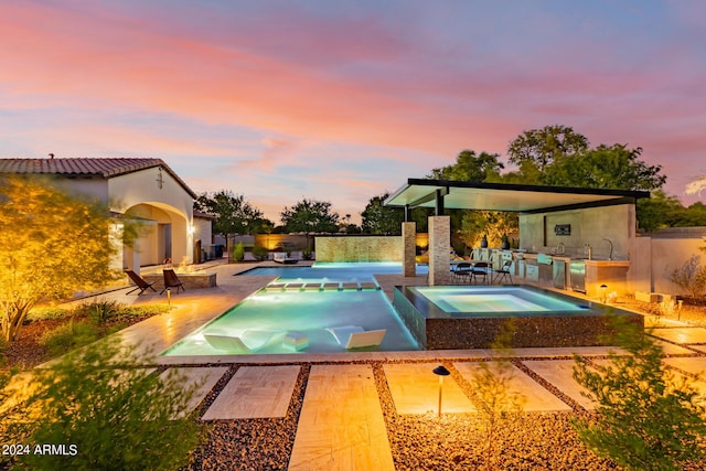 pool at dusk featuring an in ground hot tub, exterior kitchen, a patio area, and a fenced in pool