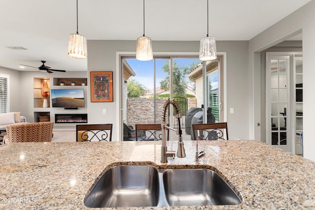 kitchen featuring a ceiling fan, light stone countertops, a sink, a glass covered fireplace, and decorative light fixtures