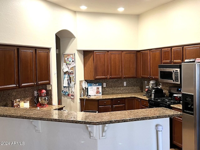 kitchen with kitchen peninsula, backsplash, stainless steel appliances, dark stone counters, and a kitchen bar