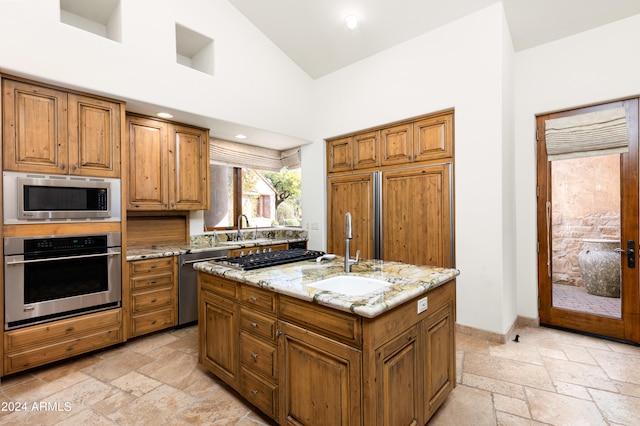 kitchen featuring an island with sink, sink, light stone counters, appliances with stainless steel finishes, and high vaulted ceiling