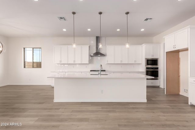 kitchen with white cabinets, a center island with sink, oven, and wall chimney range hood