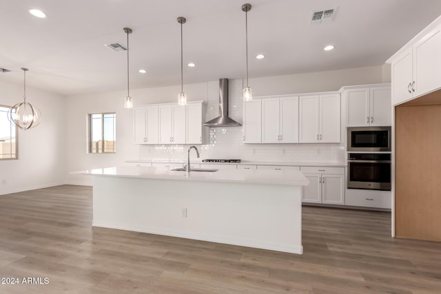 kitchen with appliances with stainless steel finishes, wall chimney exhaust hood, sink, a center island with sink, and white cabinets