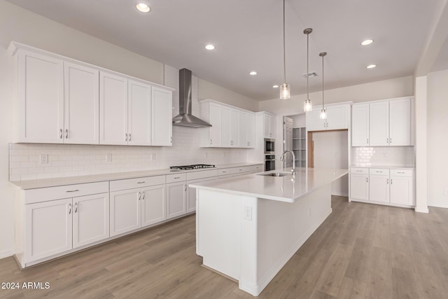 kitchen with wall chimney exhaust hood, a kitchen island with sink, sink, pendant lighting, and white cabinetry