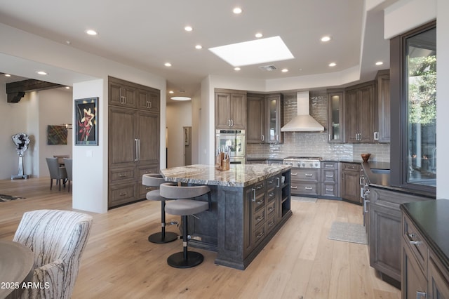 kitchen featuring wall chimney exhaust hood, a center island, stainless steel appliances, a skylight, and dark brown cabinetry