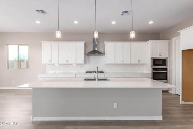 kitchen featuring appliances with stainless steel finishes, decorative light fixtures, a center island with sink, and wall chimney range hood