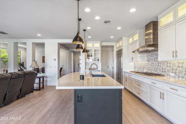 kitchen with pendant lighting, sink, stainless steel appliances, a center island with sink, and wall chimney exhaust hood