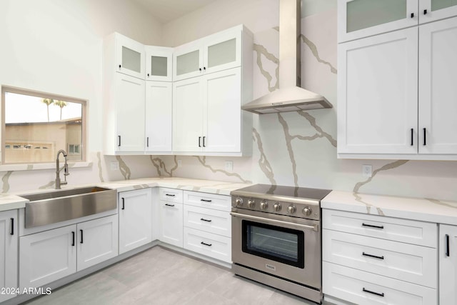 kitchen with light stone counters, wall chimney range hood, sink, white cabinets, and stainless steel stove