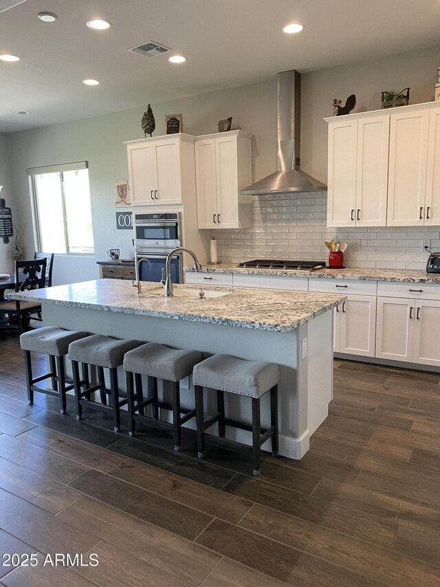 4114 Copperhead Dr, Wickenburg, AZ 85390 - kitchen featuring wall chimney range hood, stainless steel appliances, dark wood-style flooring, white cabinetry, and a breakfast bar