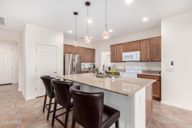 kitchen with light stone countertops, an island with sink, decorative light fixtures, white appliances, and a breakfast bar